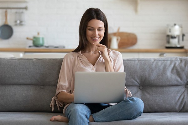 A woman smiles as she sits on her sofa looking at her laptop.