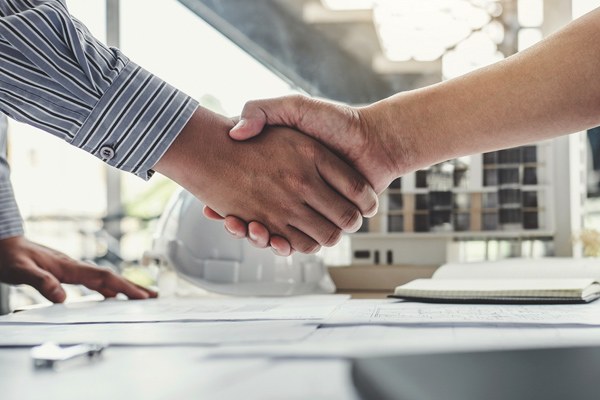 Close up of two people shaking hands in front of a hardhat on a table covered in plans.