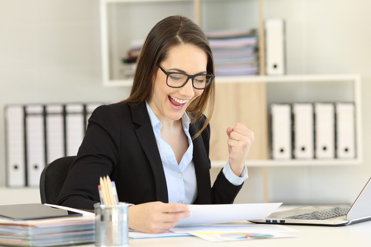 A woman in a business suit celebrates as she reads a paper at her desk.