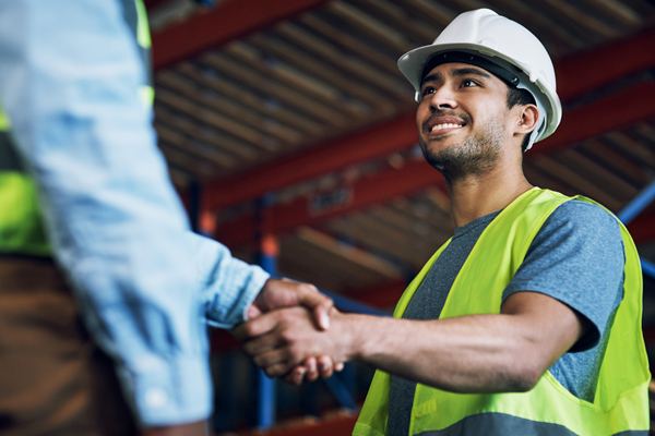 safety professional wearing a white hard hat and yellow vest and shaking hands with another person