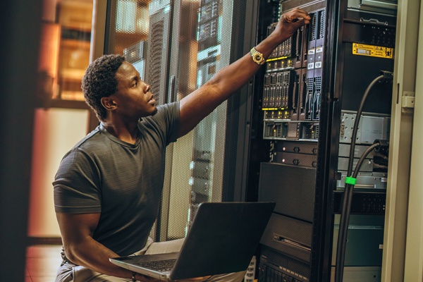 A man sits with a laptop as he checks equipment in a server room.