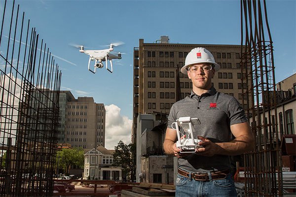 John Macahan is wearing safety glasses and helmet as he operates a drone on a construction site.
