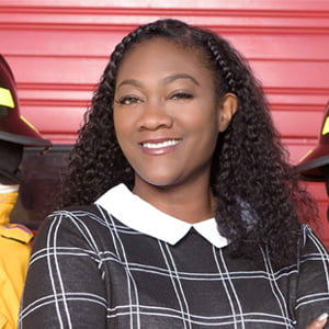 African American woman with curly shoulder length hair wearing collared blouse in front of red wall.