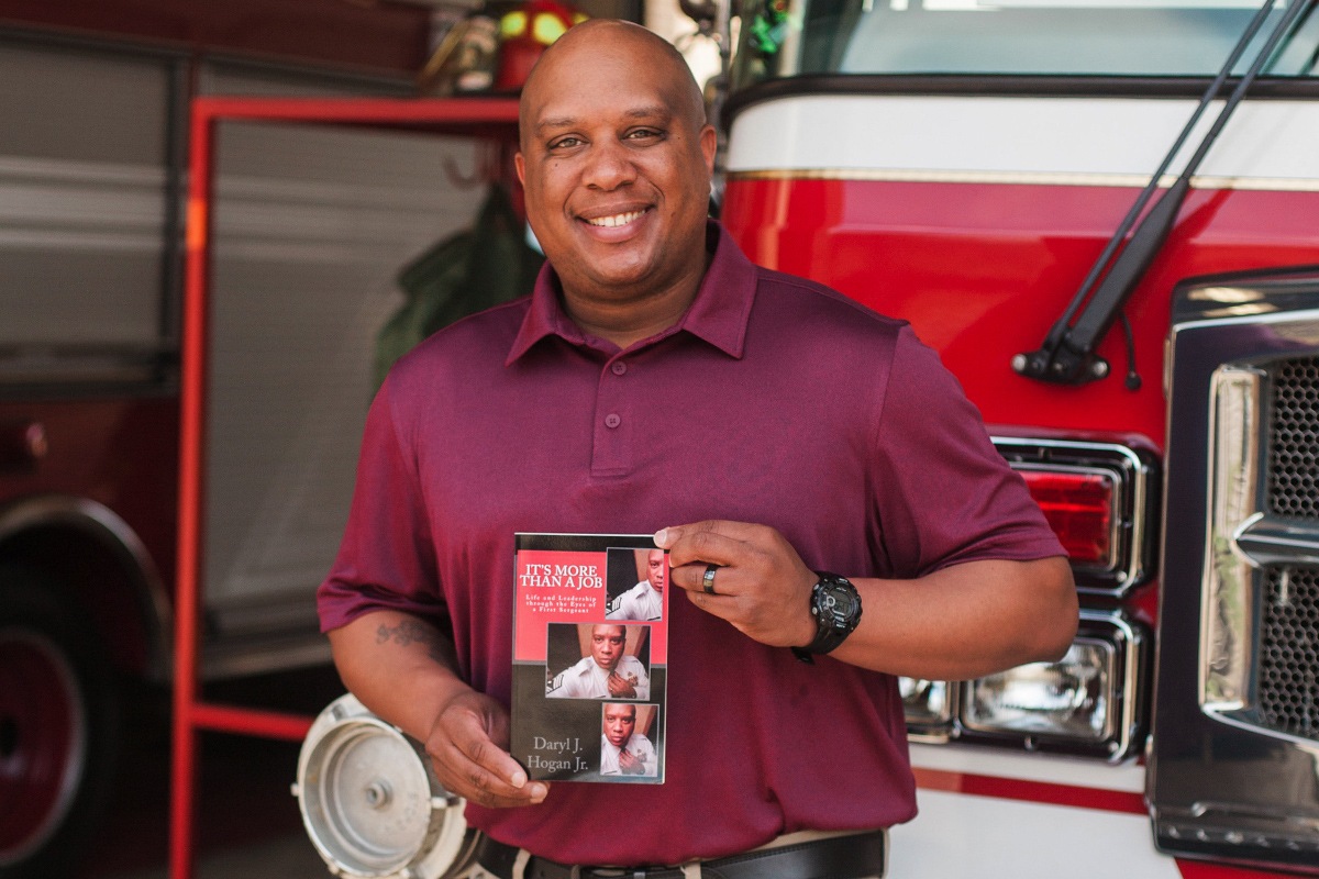 Daryl Hogan smiles in front of a fire truck as he holds his book "It's More Than a Job".