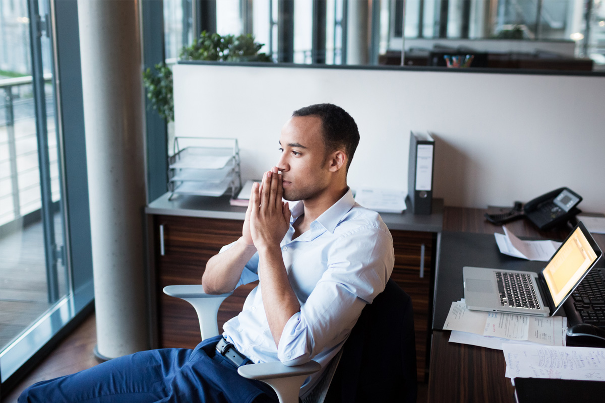 Military service member wearing civilian clothes, sitting in an office, faced away from the computer and holding hands in front of face while thinking