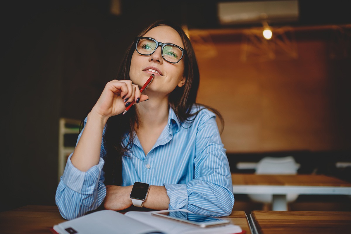 A woman sits at a desk holding a pen to her chin while looking off in the distance.