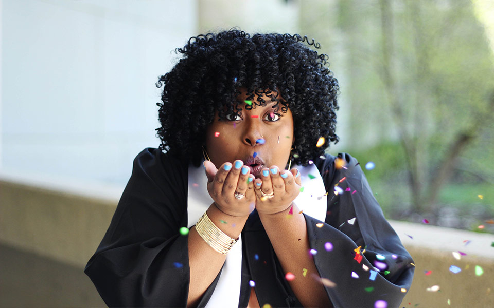 A graduate in commencement garb blows confetti towards the camera.