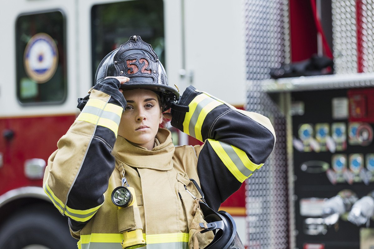 Female firefighter adjusts her helmet beside a fire truck.