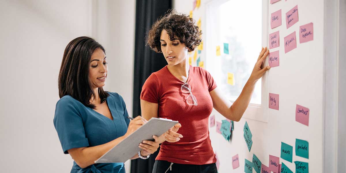 two education professionals review notes on a clipboard and sticky notes on a whiteboard