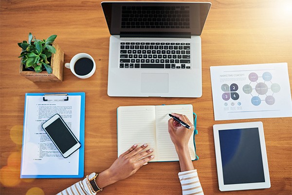 A person offscreen prepares to write at a desk surrounded by a cell phone, clipboard, laptop and tablet.