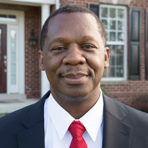 Patrick Austin is wearing a grey suit with white shirt and red tie, standing in front of brick building.