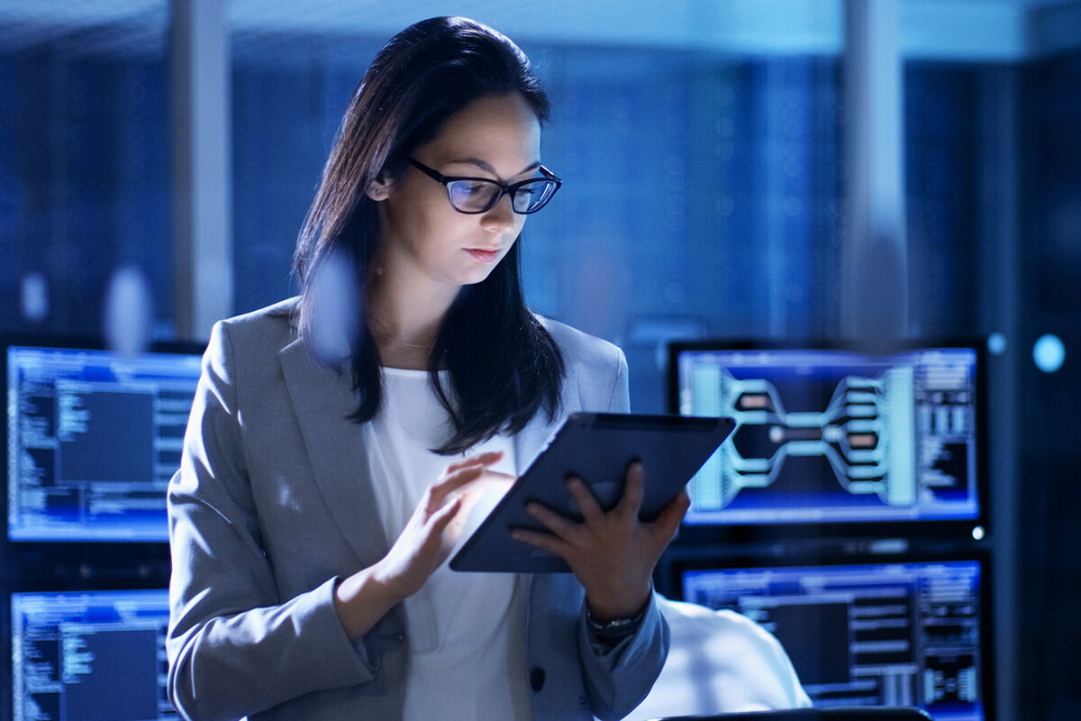 A woman in glasses and a business suit stands in a room full of computers looking intently at a tablet in her hands.