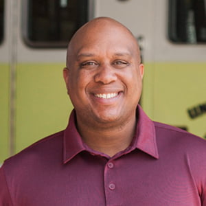 A bald, clean shaven African American man smiles. He is wearing a maroon polo shirt.