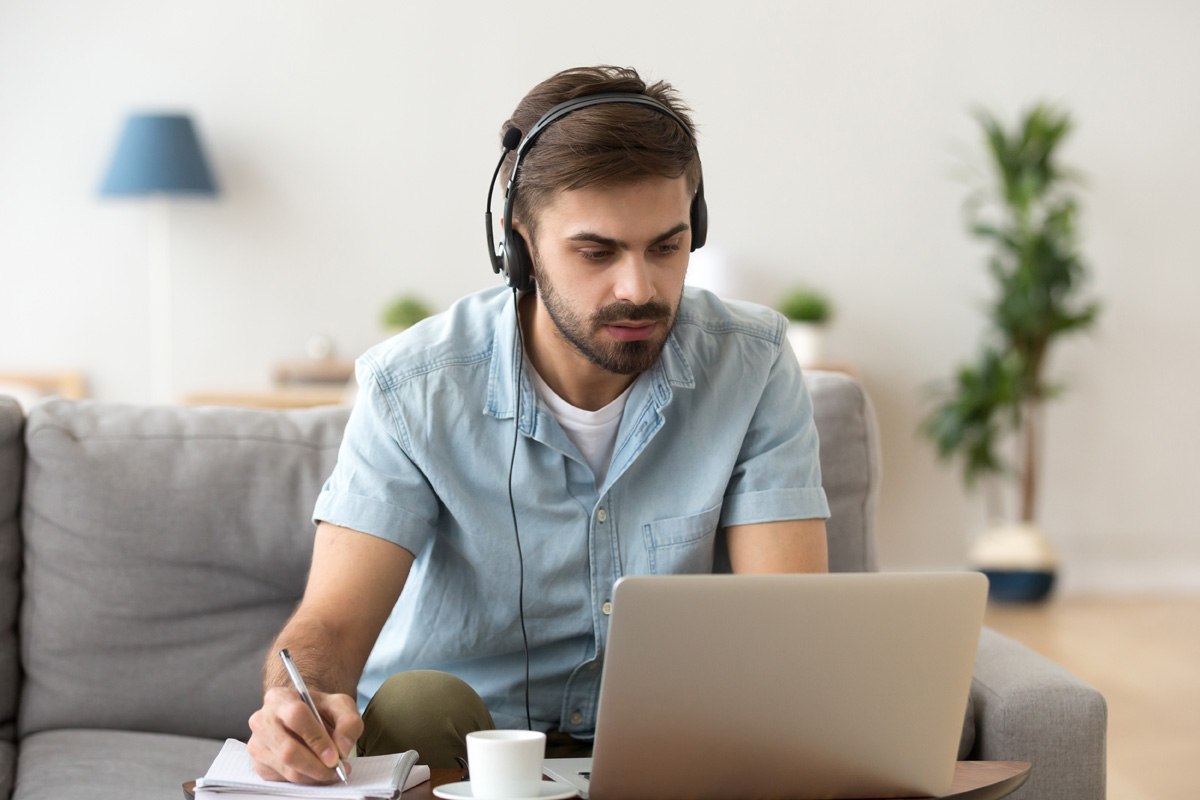 A young man wearing a headset takes notes while working at a laptop