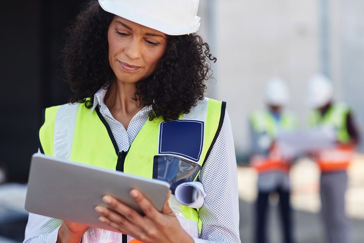 A woman in safety vest and helmet looks at a tablet.