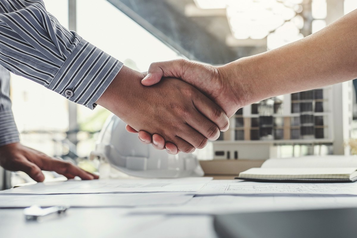 Close up of two people shaking hands in front of a hardhat on a table covered in plans.