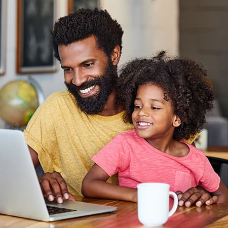 Father works on laptop as young daughter sits in his lap and looks on