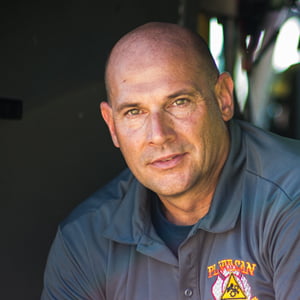 A clean shaven bald Caucasian man wearing a branded grey polo looks stoically into the camera.