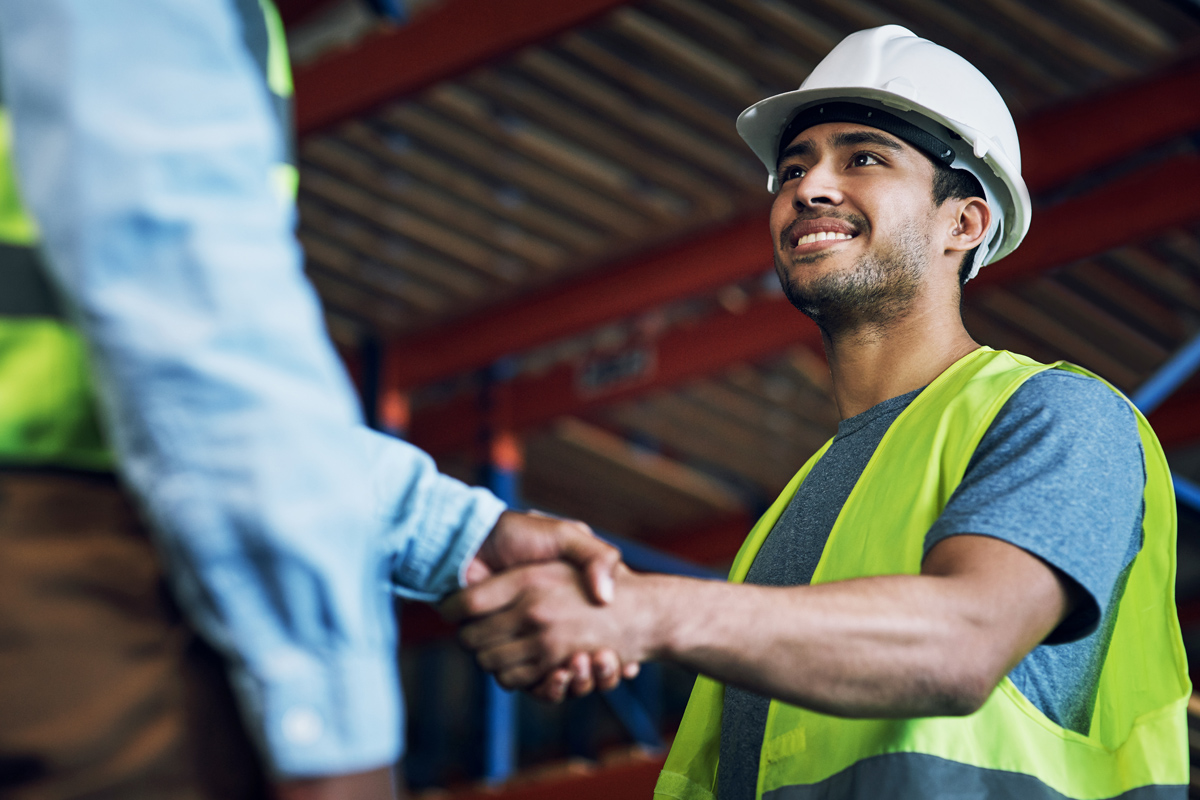 safety professional wearing a white hard hat and yellow vest and shaking hands with another person