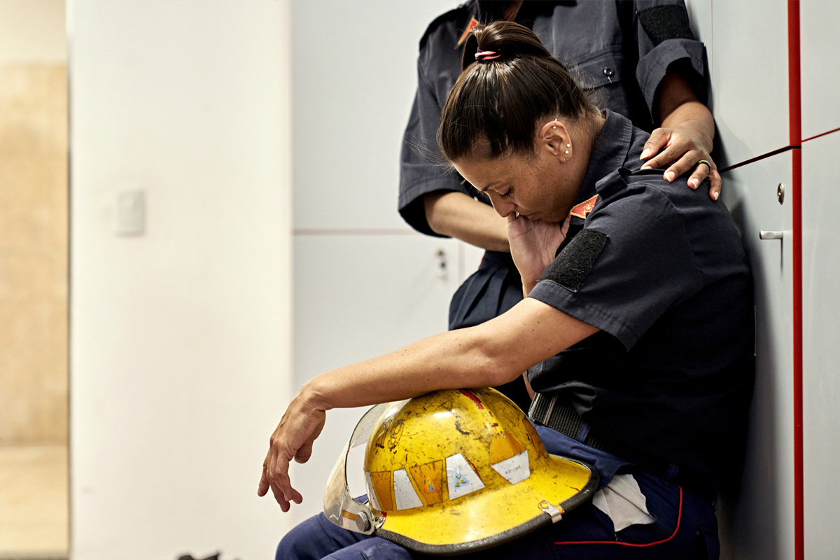 first responder sitting holding a helmet with their eyes closed while a coworker stands next to them and comforts them