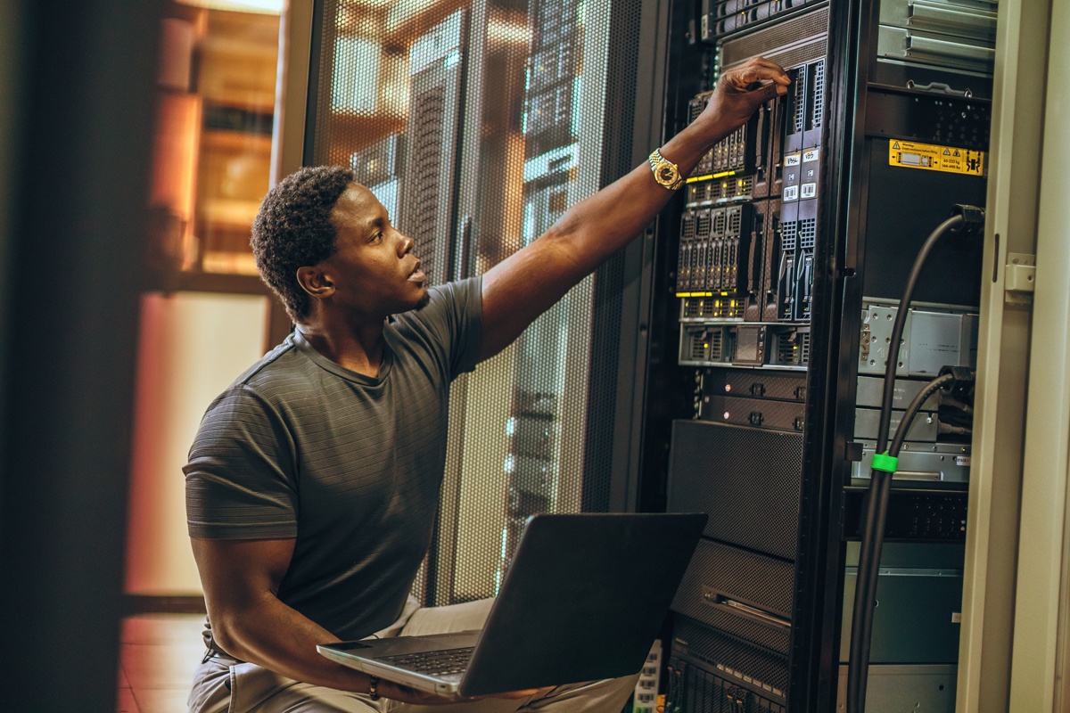 A man sits with a laptop as he checks equipment in a server room.