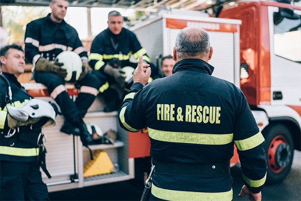 A fire & rescue leader talks to a group of firefighters sitting on and standing around a fire truck.