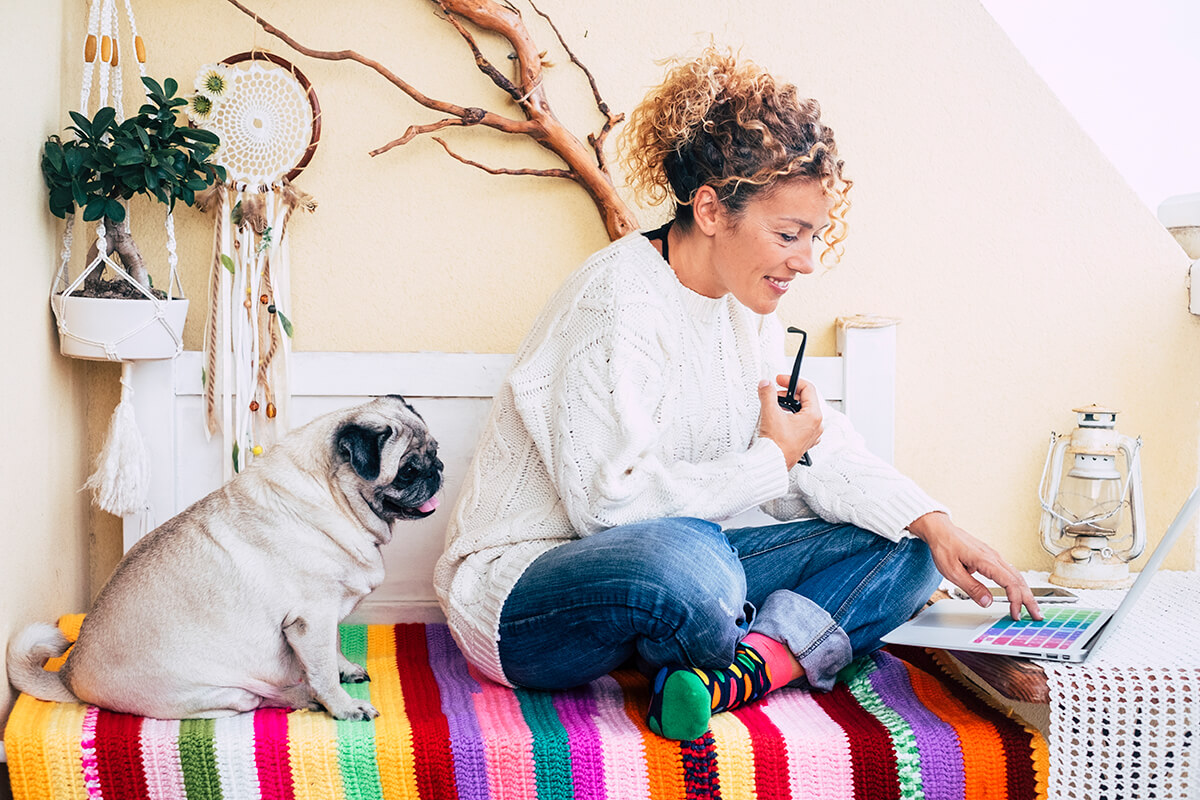 A woman sits comfortably next to her dog as she cheerfully types on a laptop.