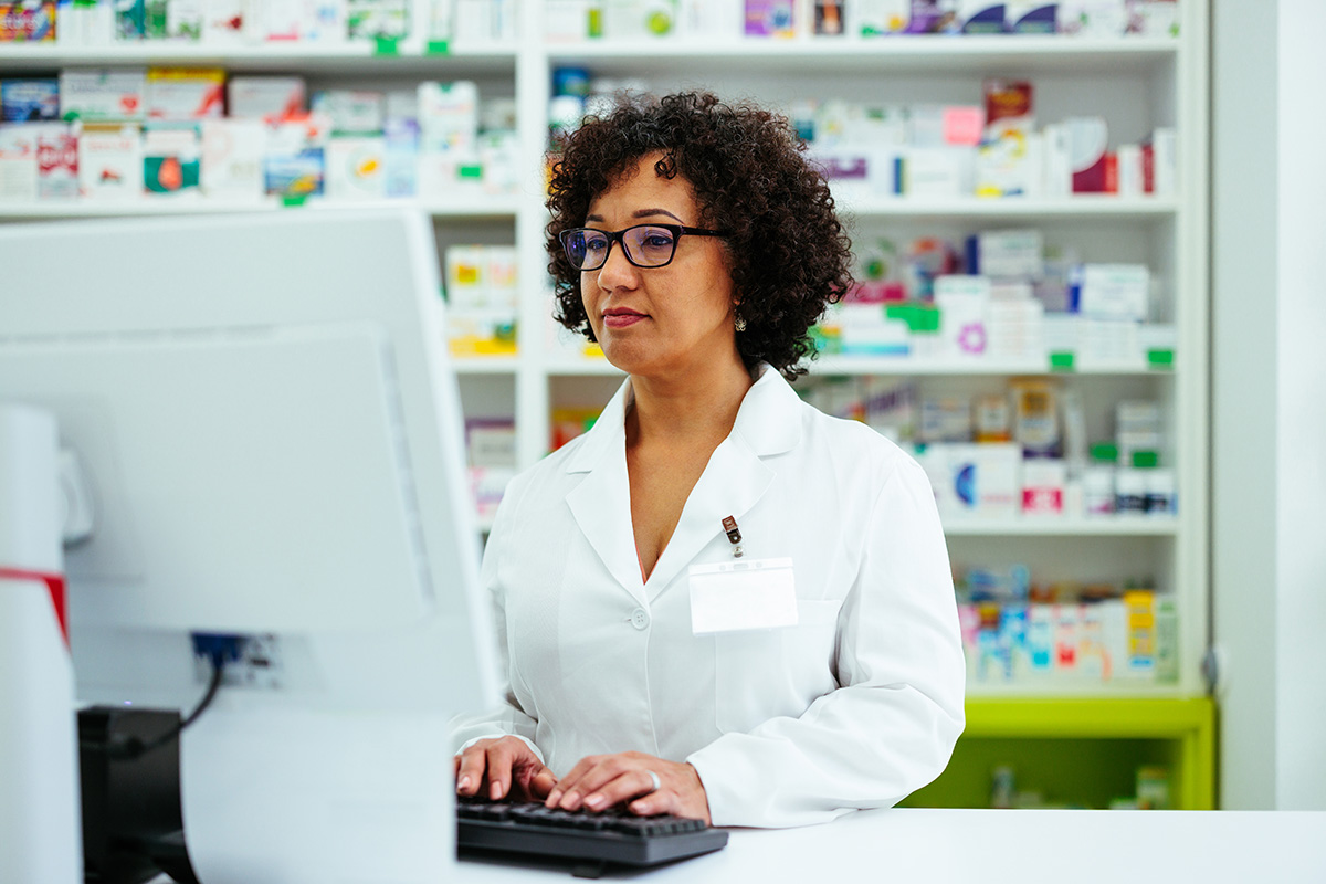 pharmacy technician working on a computer with medicines on shelves in background