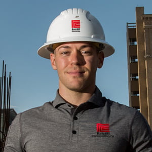 A young clean shaven Caucasian man stands proudly in a white safety helmet and branded grey polo shirt.
