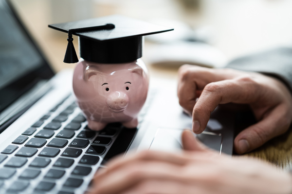closeup image of hands using a laptop while a small piggybank wearing a graduation cap rests on the keyboard