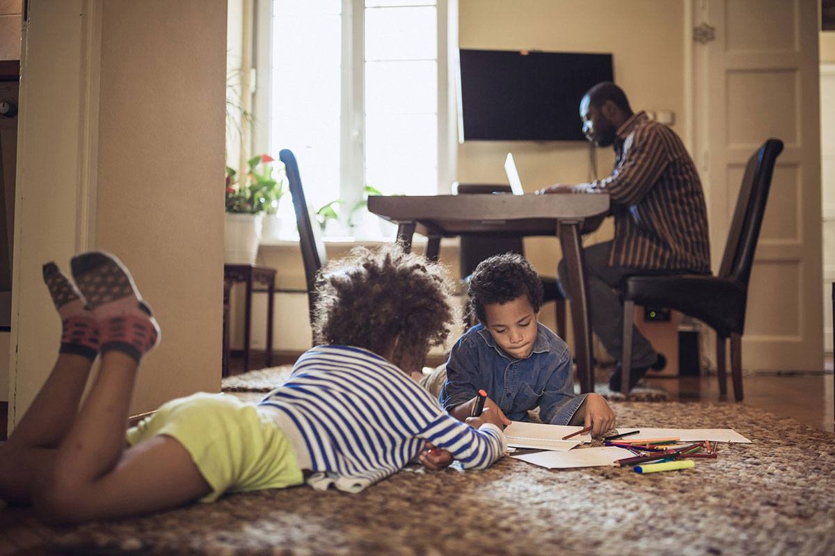 Two children lie on the floor coloring while their dad works on his laptop in the background.